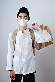 Portrait Asian Muslim man against on grey background with face mask holding box of souvenir or gifts with backpack, ready travel