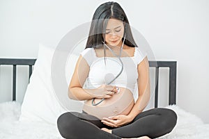 Portrait of Asian pregnant mother use stethoscope to listen sound of baby in her womb during sit on bed with morning light.