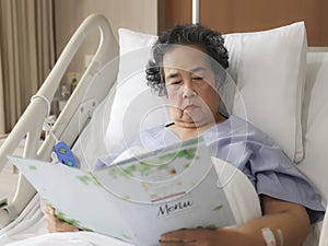 Asian senior female patient  sitting and reading food menu in hospital bed, ordering food. Elderly health concept