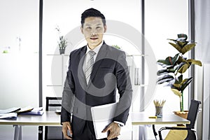 Portrait of Asian smiling businessman in suit holding laptop computer at desk in the office, man in workplace, doing business