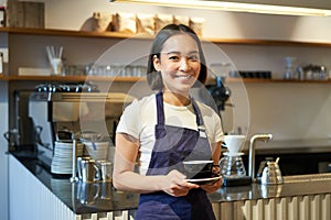Portrait of asian smiling female barista, cafe waitress holding cup of coffee, serving clients, taking order to the