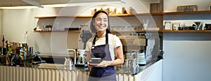 Portrait of asian smiling female barista, cafe waitress holding cup of coffee, serving clients, taking order to the