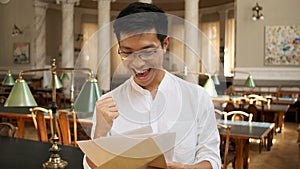 Portrait of asian student joyfully opening envelope with exam results in university library. Young attractive guy
