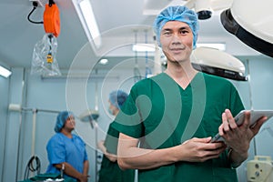 Portrait of Asian surgeon with medical mask standing and holding a tablet in operation theater at a hospital. Team of Professional