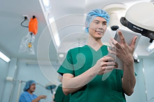 Portrait of Asian surgeon with medical mask standing and holding a tablet in operation theater at a hospital. Team of Professional