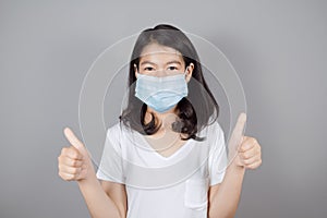 Portrait of Asian teenager  in medical face mask to protect Covid-19 Coronavirus wears white t-shirt showing thumb up on gray