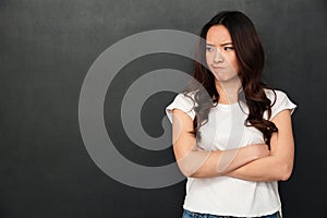 Portrait of asian woman in casual t-shirt standing with hands folded and looking aside with resentment, isolated over dark gray b