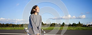 Portrait of asian woman with longboard. Korean girl skating, holding skateboard in hands, posing on road, smiling and