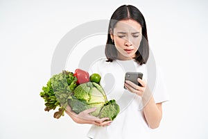 Portrait of asian woman looks worries at smartphone screen, holds vegetables, orders food online, white background