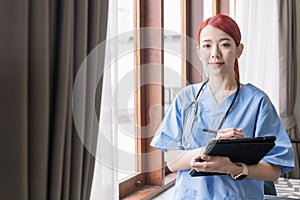 Portrait of Asian woman nurse wearing medical scrubs, stethoscope and holding the tablet in the house of patient. Caregiver visit