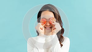 Portrait of asian woman smiling and holding tomato slices on eyes on blue