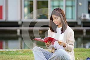 Portrait of Asian woman university student aitting on grass in campus looking happy and reading a book in park