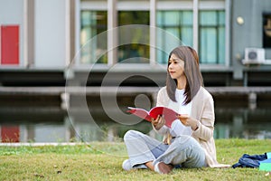 Portrait of Asian woman university student aitting on grass in campus looking happy and reading a book in park