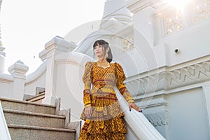 Portrait Asian woman wearing a yellow Thai patterned dress standing on a stair at a white Buddhis pagoda while traveling in Asia