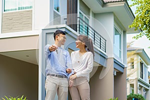 Portrait of Asian young couple standing and hugging together and holding house key looking happy in front of their new house to