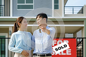 Portrait of Asian young couple standing and hugging together and holding house key looking happy in front of their new house to