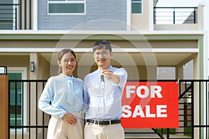 Portrait of Asian young couple standing and hugging together and holding house key looking happy in front of their new house to