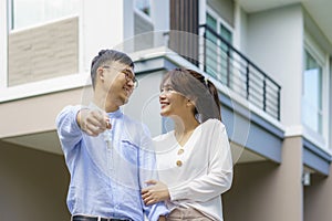 Portrait of Asian young couple standing and hugging together and holding house key looking happy in front of their new house to