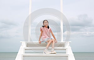 Portrait of Asian young girl child looking camera with smiling while sitting on white stair at seaside