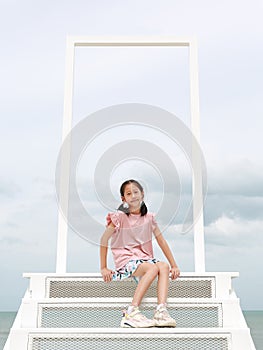 Portrait of Asian young girl child looking camera with smiling while sitting on white stair at seaside