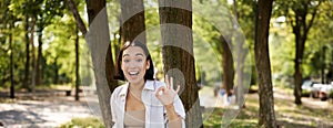 Portrait of asian young woman, student doing homework, working in park, sitting beside tree with laptop and showing okay