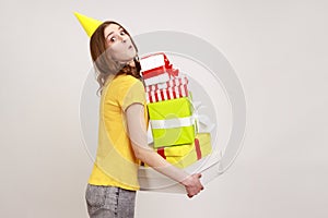 Portrait of astonished brown haired young woman with party cone holding many present boxes on