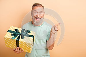 Portrait of astonished cheerful aged man hold desirable giftbox indicate finger empty space proposition isolated on