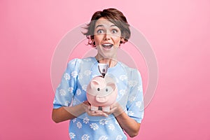 Portrait of astonished positive girl open mouth arms hold money bank pig dollar bills isolated on pink color background