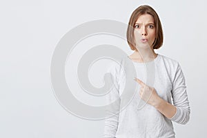 Portrait of astonished pretty young woman in longsleeve feeling amazed and pointing to the side on copyspace isolated over white