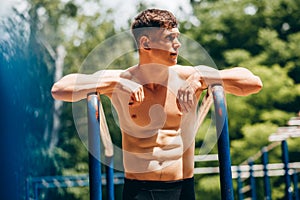 Portrait of athlete young man resting after exercises on horizontal crossbar at sportsground. Handsome muscular shirtless male