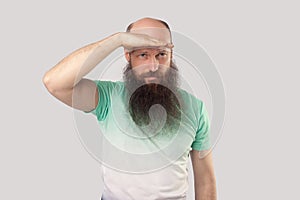 Portrait of attentive middle aged bald man with long beard in light green t-shirt standing with hand on forehead and looking far