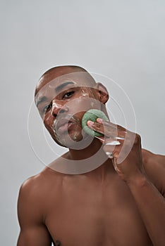 Portrait of attractive african american young man using cleansing sponge while washing his face, standing isolated over