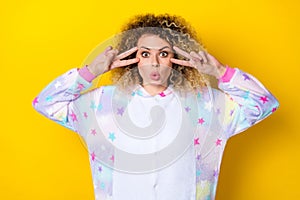 Portrait of attractive amazed funny wavy-haired girl in pajama showing v-sign near eye  over bright yellow color