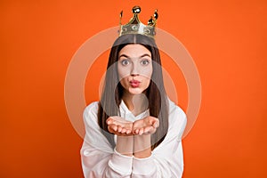 Portrait of attractive amorous girl wearing crown sending air kiss isolated over bright orange color background