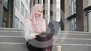 Portrait of attractive arabian woman in elegant clothes and hijab sitting on stairs with modern laptop on knees