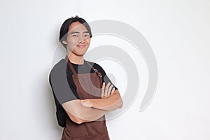 Portrait of attractive Asian barista man in brown apron keeping arms crossed and looking at camera. Advertising concept. Isolated