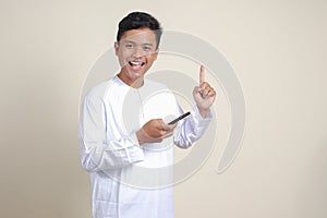 Portrait of attractive Asian muslim man in white shirt holding mobile phone with smiling expression on face while pointing finger