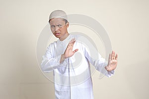 Portrait of attractive Asian muslim man in white shirt with skullcap forming a hand gesture to avoid something or bad things.