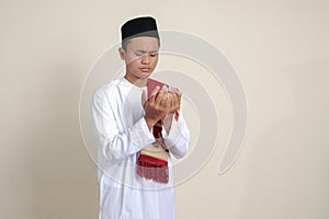 Portrait of attractive Asian muslim man in white shirt with skullcap praying earnestly with his hands raised. Isolated image on