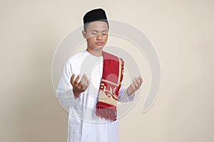 Portrait of attractive Asian muslim man in white shirt with skullcap praying earnestly with his hands raised. Isolated image on