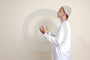 Portrait of attractive Asian muslim man in white shirt with skullcap praying earnestly with his hands raised. Isolated image on