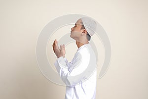 Portrait of attractive Asian muslim man in white shirt with skullcap praying earnestly with his hands raised. Isolated image on