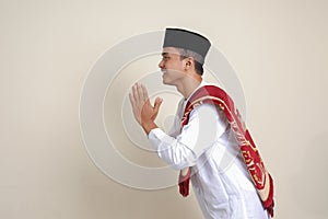 Portrait of attractive Asian muslim man in white shirt with skullcap showing apologize and welcome hand gesture. Isolated image on