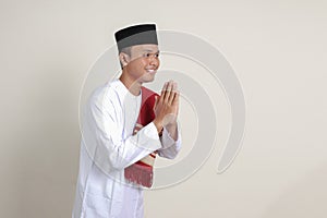 Portrait of attractive Asian muslim man in white shirt with skullcap showing apologize and welcome hand gesture. Isolated image on