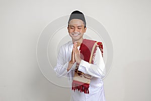 Portrait of attractive Asian muslim man in white shirt with skullcap showing apologize and welcome hand gesture. Isolated image on