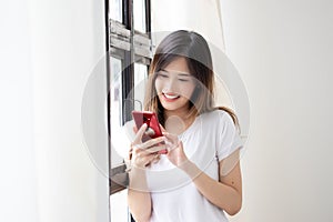 Portrait of attractive asian woman in casual clothes is using a smart phone and smiling while standing near the window, holding