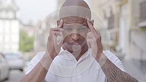 Portrait of attractive bald middle eastern man standing in the city street massaging his forehead with fingers. The guy