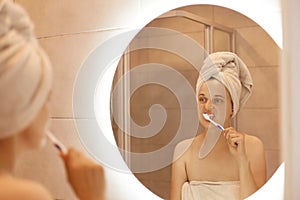 Portrait of attractive beautiful woman with towel on her head standing in front of the mirror in the bathroom and brushing her