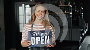 Portrait of attractive blond waitress holding open sign in new cafe smiling