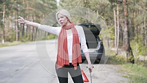 Portrait of attractive blond woman with wrench in hand hitchhiking in front of broken car with open trunk. Beautiful